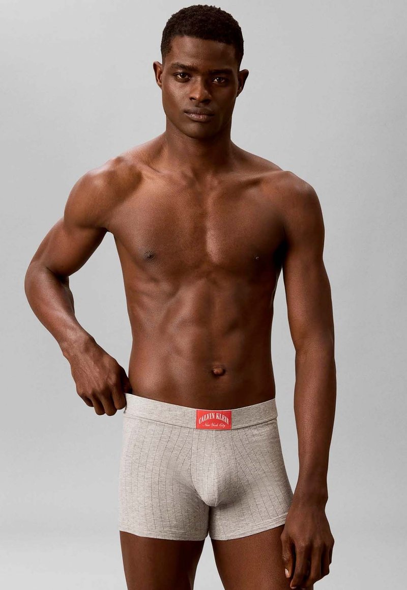 Homme torse nu avec une silhouette musclée portant un boxer gris clair Calvin Klein, ajustant la ceinture avec la main gauche sur un fond gris uni.
