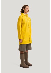Kvinna med lockigt hår i en gul regnrock, grå knälånga shorts och bruna gummistövlar som står mot en ljus bakgrund och vänder sig åt höger.