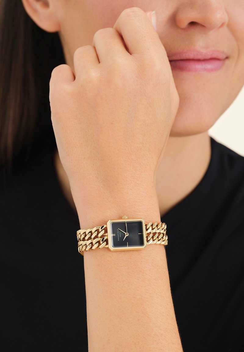 Pols met een rechthoekig zwart-gezichts goudkleurig horloge met een dubbele goudkleurige schakelarmband, gedragen door een persoon in een zwart shirt.