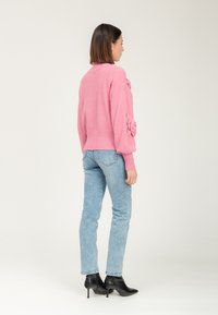 Donna in jeans azzurri e maglione rosa a maglia con dettagli floreali sulle maniche, che indossa stivaletti neri con tacco, vista di lato.