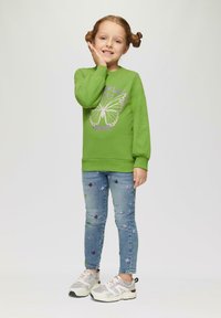 Groene sweatshirt met vlinderprint, gecombineerd met lichtblauwe jeans met bloem- borduursel en grijze sneakers met roze accenten.