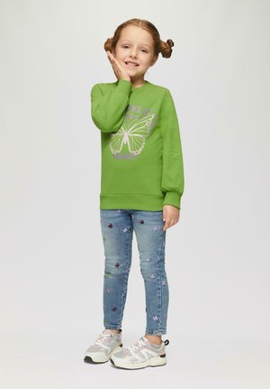 Groene sweatshirt met vlinderprint, gecombineerd met lichtblauwe jeans met bloem- borduursel en grijze sneakers met roze accenten.