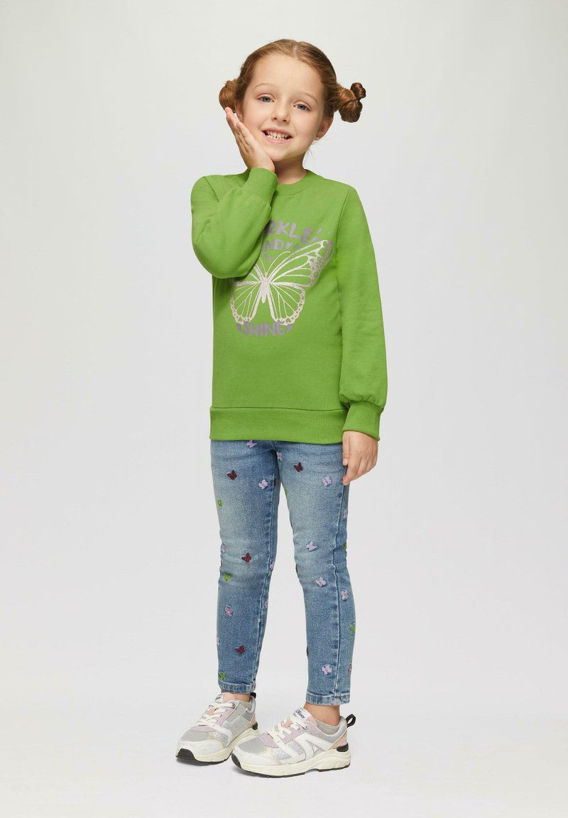 Groene sweatshirt met vlinderprint, gecombineerd met lichtblauwe jeans met bloem- borduursel en grijze sneakers met roze accenten.