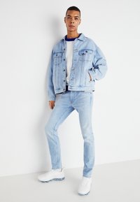 Chaqueta de mezclilla azul claro con dos bolsillos en el pecho, llevada sobre una camiseta blanca. Jeans azul claro y zapatillas blancas completan el atuendo.