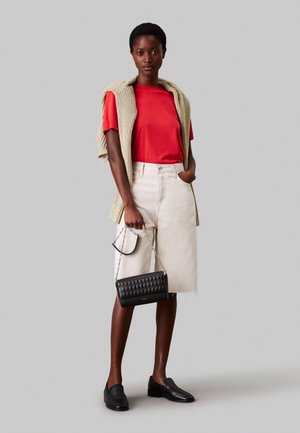 Kobieta ubrana w czerwony t-shirt, beżową spódnicę do kolan, sweter na ramionach, czarne mokasyny, trzyma czarną pikowaną torebkę na łańcuszku.