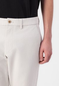 Pantalon beige clair en tissu doux, avec fermeture à bouton, poches latérales et coupe ajustée avec une texture subtile.