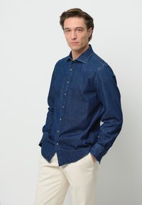 Camicia in denim scuro con colletto a punta, maniche lunghe e polsini con bottone. Indossata con pantaloni color crema. Design semplice, senza fantasie.