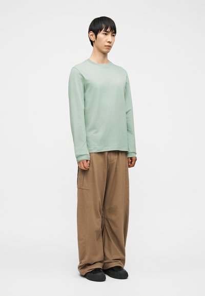 Νεαρός άνδρας στέκεται φορώντας ανοιχτοπράσινο πουκάμισο με μακριά μανίκια, χαλαρά καφέ παντελόνια και μαύρα παπούτσια, μπροστά σε λευκό, απλό φόντο.