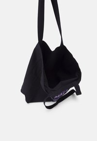 Sac cabas noir en tissu robuste. Il comporte de larges bretelles et une ouverture en haut, avec un logo violet imprimé sur un côté.