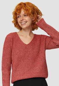 Roter, gestrickter Pullover mit V-Ausschnitt, locker sitzend und langen Ärmeln, die ein dezentes Strukturmuster aufweisen. Schwarze, hochtaillierte Hose.