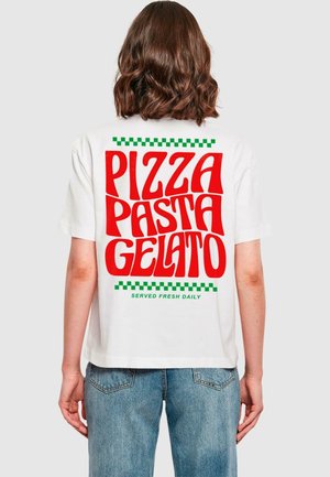 Osoba z brązowymi włosami do ramion nosi białą koszulkę z czerwonym napisem „PIZZA PASTA GELATO” i zielonymi kratkowymi akcentami oraz niebieskie dżinsy.