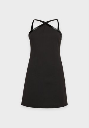 Une femme porte une mini-robe noire à bretelles croisées et des chaussures noires à talons hauts sur un fond clair et uni.