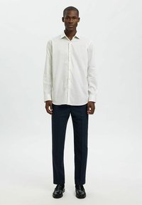 Mann steht da und trägt ein weißes, knopfbares Langarmhemd, dunkle formelle Hosen und schwarze Schuhe vor einem einfarbigen Hintergrund.