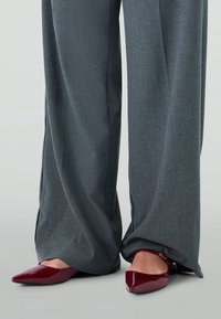 Ballerines en cuir verni bordeaux avec bouts pointus, associées à un pantalon large gris avec une texture subtile et des fentes sur les côtés.
