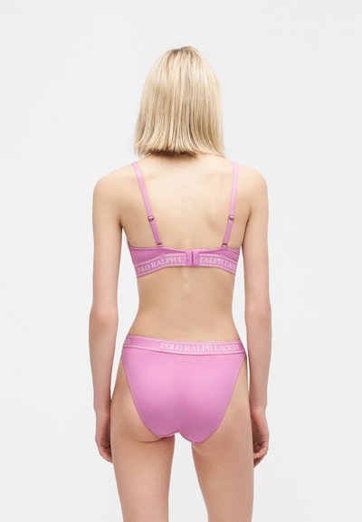 Γυναίκα που στέκεται με την πλάτη προς τα εμπρός, φορώντας ροζ σουτιέν Polo Ralph Lauren και matching σλιπ, σε ένα απλό ανοιχτό φόντο.