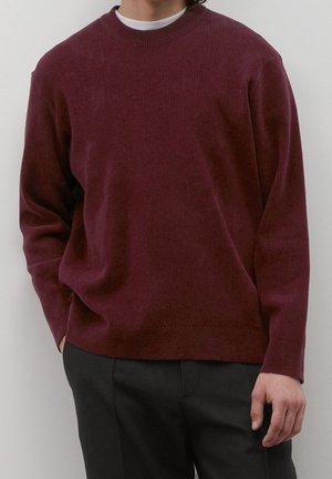 Person trägt einen dunkelroten Rippenpullover über einem weißen Unterhemd und schwarze Hosen, steht vor einem hellen Hintergrund.