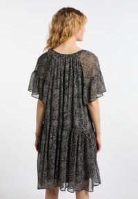 Musta kuvioitu mekko, jossa on löysä, virtaava malli, läpinäkyvä kangas, lyhyet kellomaiset hihat ja kerroksellinen laskostettu vyötärö. Takakatselma esittelee koottua yksityiskohtaa.