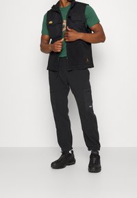 Černá vesta s předními kapsami přes zelené tričko. Černé cargo kalhoty s texturovanými panely a černé tenisky. Jednoduchý, funkční design.