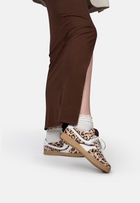 Sneaker con stampa leopardata e dettagli bianchi, suola beige, abbinate a calze bianche con volant, indossate con un capo marrone a spacco laterale.