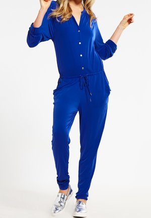 Femme portant une combinaison bleu roi à manches longues avec des boutons sur le devant et un cordon à la taille, associée à des chaussures argentées métalliques sans lacets.
