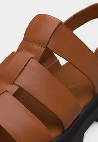 Κοντινή λήψη ενός καφέ δερμάτινου σανδάλι με φαρδιές λουριά και μαύρη σόλα, που δείχνει λεπτομερή ραφή και λεία υφή.