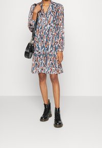 Tištěné šaty s dlouhým rukávem a výstřihem do V, s vícevrstvou sukní v odstínech modré, oranžové a šedé. Kombinované s černými šněrovacími botami a černou kabelkou.