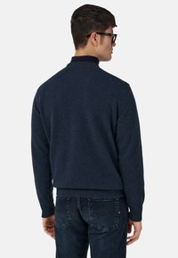 Granatowy dzianinowy sweter z kołnierzem stójką, prążkowanymi mankietami i dołem; w zestawieniu z ciemnymi jeansami. Widok od tyłu ukazuje proste, czyste linie.