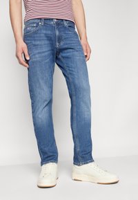 Jeans de mezclilla azul con corte recto, diseño de cinco bolsillos y un ligero desgaste. Combinados con zapatillas blancas y una camiseta a rayas.