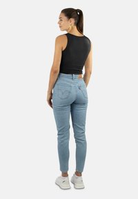 Frau mit Pferdeschwanz, die ein schwarzes ärmelloses Oberteil, hellblaue hochtaillierte Jeans, weiße Turnschuhe und goldene Blattohrringe trägt, vor einem weißen Hintergrund steht.
