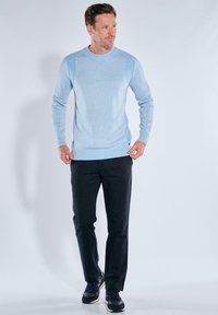 Hellblauer Strickpullover mit geribbelter Textur an den Schultern und dem Saum, kombiniert mit dunklen Hosen und schwarzen Sneakers vor einem neutralen Hintergrund.