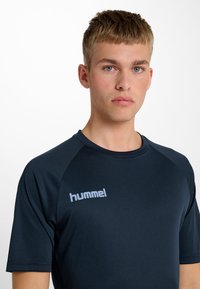 Sportowa koszulka w kolorze granatowym z krótkim rękawem i okrągłym dekoltem, z jasnoszarym logo "hummel" na lewej piersi. Gładka, dopasowana struktura.