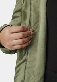 Chaqueta verde oliva con forro de vellón texturizado, que presenta un cierre de cremallera y un detalle de velcro cerca de la apertura. Mano tocando la tela.
