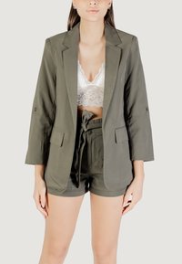 Blazer verde oliva con maniche lunghe, abbinato a un reggiseno in pizzo bianco e pantaloni corti verdi oliva coordinati con vita legata.