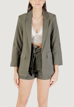 Blazer verde oliva con maniche lunghe, abbinato a un reggiseno in pizzo bianco e pantaloni corti verdi oliva coordinati con vita legata.