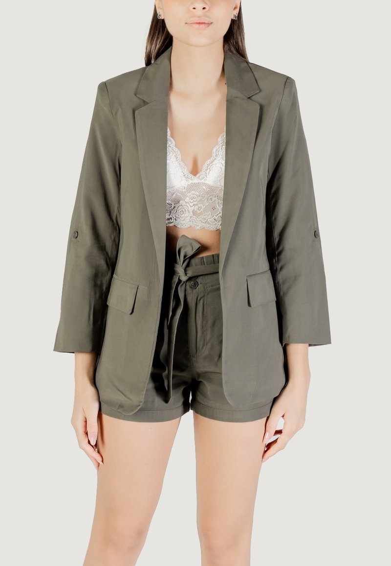 Blazer verde oliva con maniche lunghe, abbinato a un reggiseno in pizzo bianco e pantaloni corti verdi oliva coordinati con vita legata.