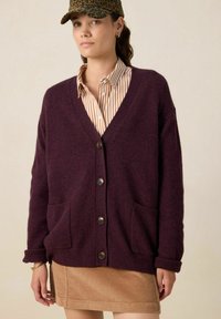 Cardigan en tricot bordeaux avec un col en V, cinq boutons, deux poches avant et des poignets côtelés. Porté par-dessus une chemise rayée et une jupe en velours côtelé.