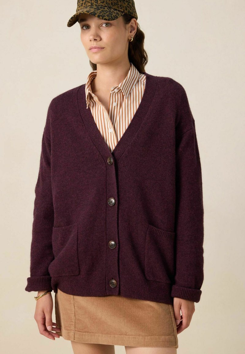 Cardigan en tricot bordeaux avec un col en V, cinq boutons, deux poches avant et des poignets côtelés. Porté par-dessus une chemise rayée et une jupe en velours côtelé.