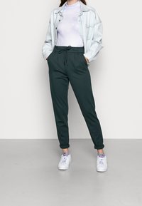 Dark green jogger pants with a drawstring waist, tapered ankles, paired with a light blue denim jacket and white sneakers.