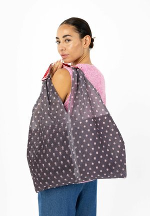 Femme aux cheveux foncés attachés, portant un haut rose sans manches et un pantalon bleu, portant un grand sac fourre-tout en filet noir avec des pois roses sur l'épaule.