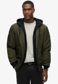 Giacca bomber verde oliva con fodera nera a cappuccio, caratterizzata da una chiusura con zip, tasche laterali e polsini a costine. Indossata sopra una maglietta grigia.