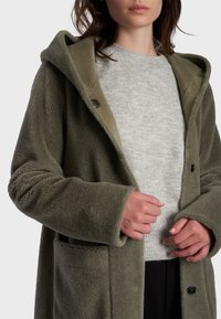 Cappotto in pile verde oliva con cappuccio, caratterizzato da una texture morbida, tasche frontali e bottoni scuri. Indossato sopra un maglione grigio chiaro.