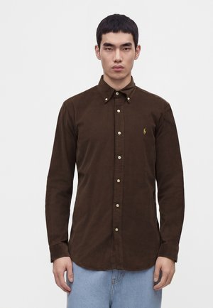 Homme portant une chemise marron à boutons avec col et un jean bleu clair, debout devant un fond blanc uni.