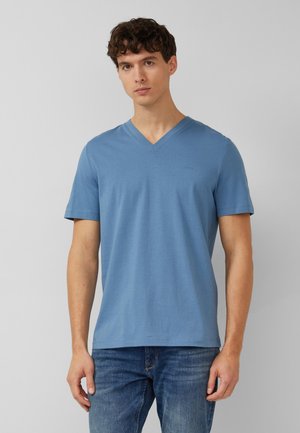 Man met kort krullend haar die een effen blauw T-shirt met V-hals en korte mouwen en een blauwe spijkerbroek draagt, staand tegen een neutrale achtergrond.