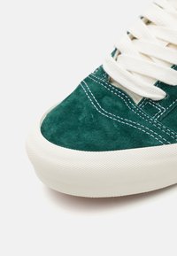 Zapatillas de ante verde con suela de goma blanca, superficie texturizada, costuras en contraste y cordones blancos planos, destacando un diseño de perfil bajo.