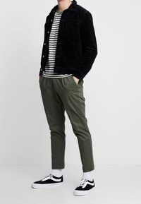 Schwarze Cord-Jacke über einem grau-weißen gestreiften T-Shirt, kombiniert mit olivgrünen, schmal zulaufenden Hosen und schwarz-weißen Sneakers.