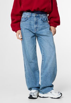 Persona que lleva vaqueros holgados de color azul claro, zapatillas blancas y una camiseta de manga larga roja sobre un fondo liso.