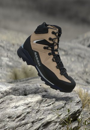 Bota de senderismo Mammut marrón y negra con suela resistente, flotando sobre un terreno rocoso con hierba escasa y pequeñas flores blancas.