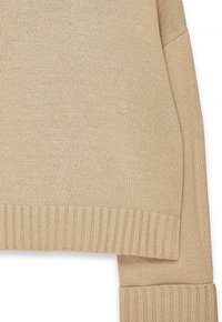Beige stickad tröja med snibbade fåll- och mudddetaljer samt en finstickad textur på en vit bakgrund.
