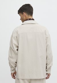 Beige kordbársony ing hosszú ujjakkal, hegyes gallérral és gombos mandzsettával. A vízszintes bordázott textúra vizuális érdekességet ad.