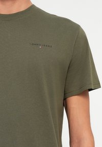 Detailní záběr na osobu v olivově zeleném tričku Tommy Jeans s krátkým rukávem a malým logem na levé hrudi.